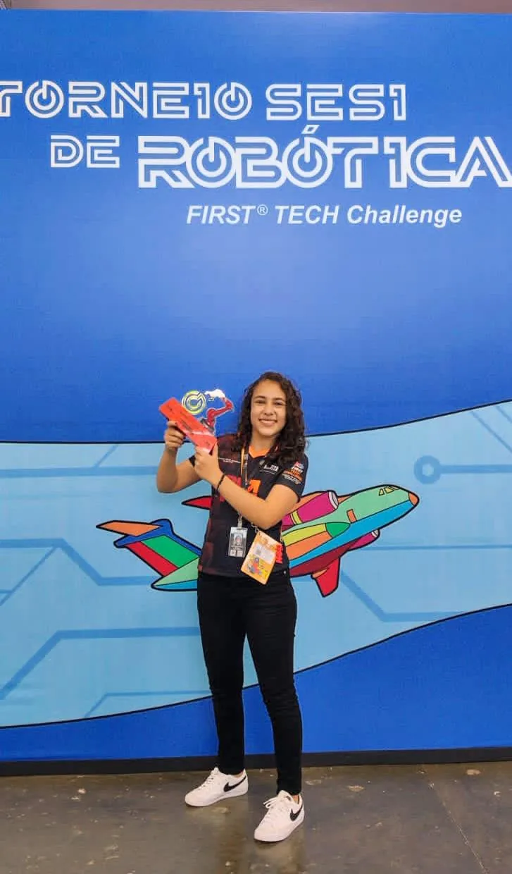 Maria Fernanda segurando troféu diante do painel do Torneio SESI de Robótica.